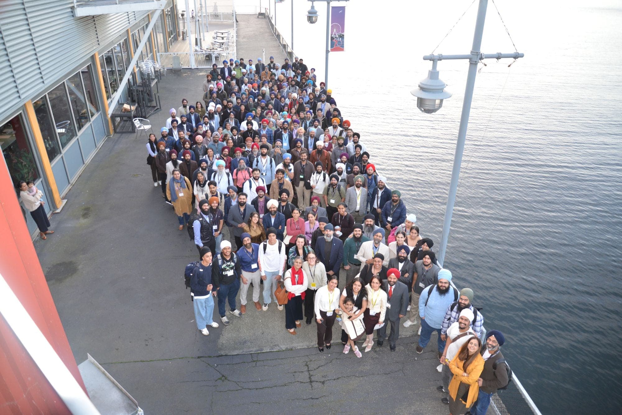 The Sarbat sangat gathered for a group photo on the pier beside the water, seen from above.