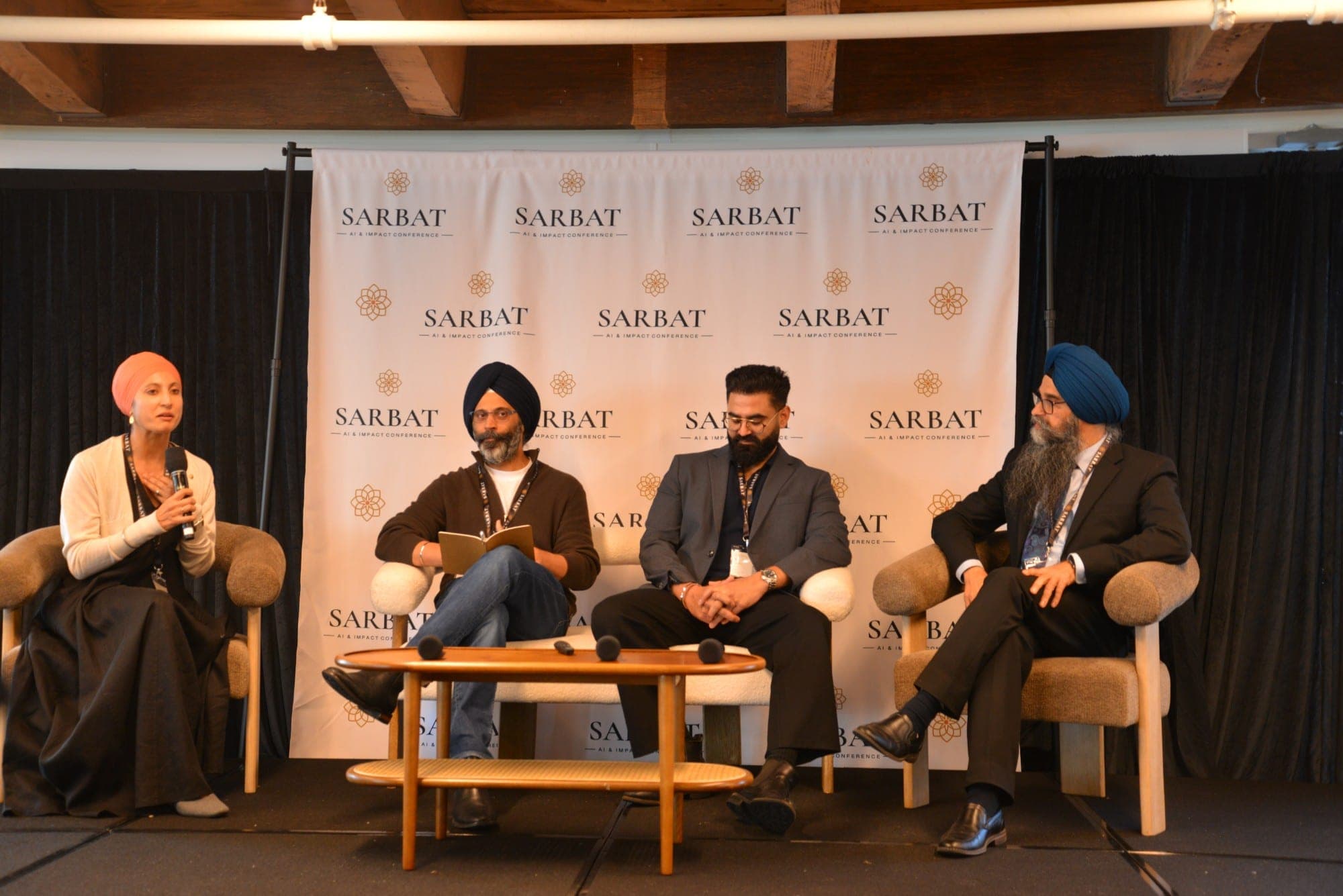 Four panelists in armchairs on stage in front of the Sarbat backdrop, one speaking into a microphone.