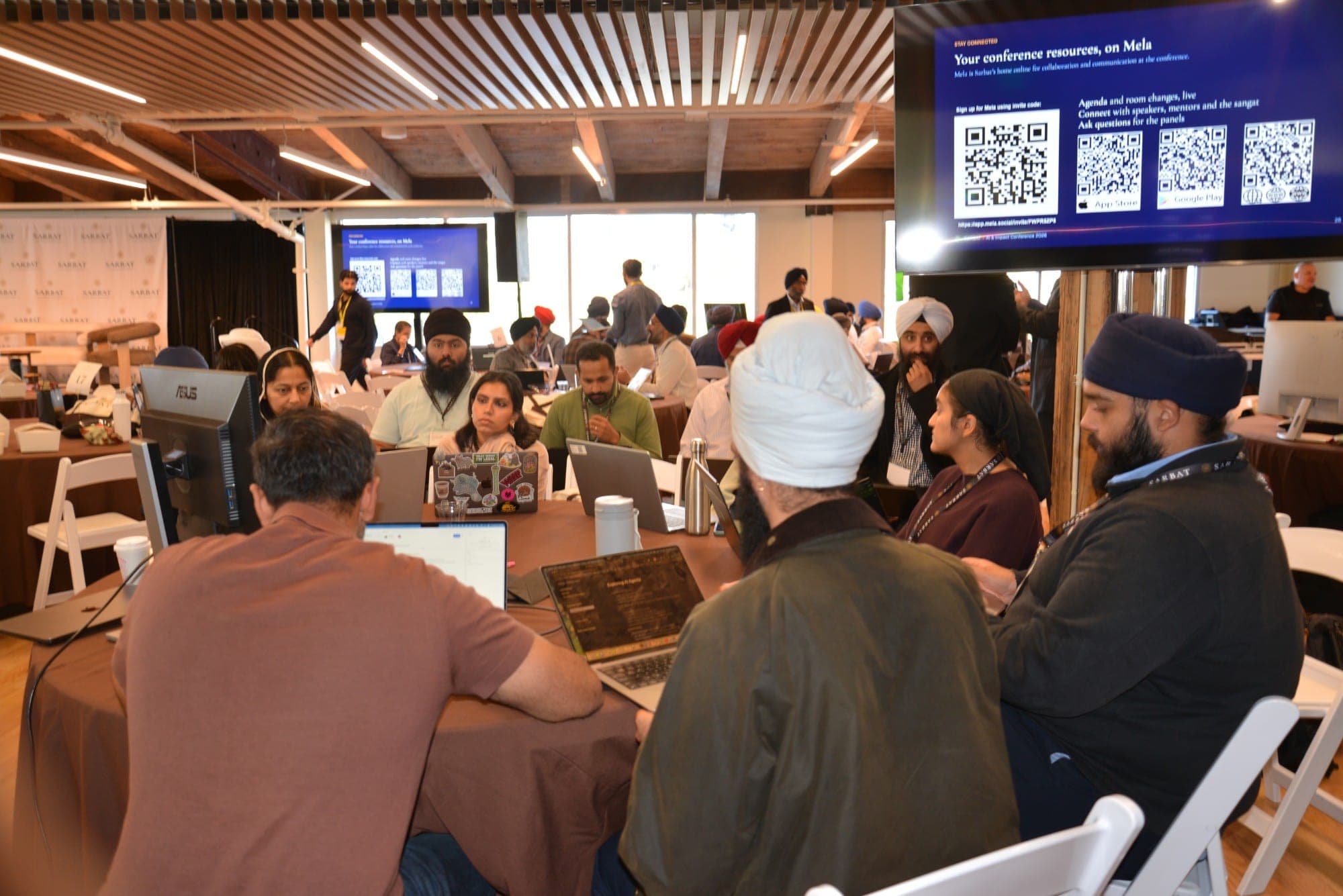 Attendees working on laptops around a round table, with a screen of conference resources overhead and more tables beyond.
